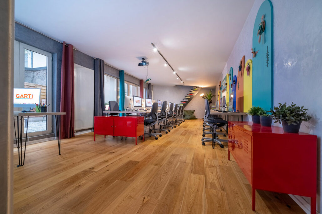 Pièce informatique avec du parquet marron, des rideaux de couleurs, des bureaux et chaises gris et des ordinateurs mac.